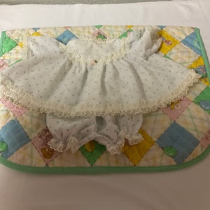 May include: A white baby doll dress with a floral print and lace trim. The dress is on a colourful quilted fabric background.