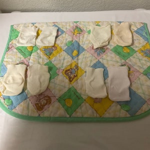 May include: A collection of eight pairs of off-white baby socks arranged on a colorful, quilted play mat. The mat features a patchwork design with pastel squares and cartoon illustrations, edged with a green border.