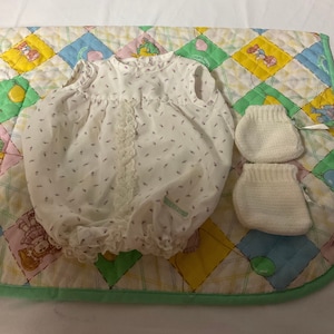 May include: A white baby doll outfit with a floral print and lace trim. The outfit is on a quilted blanket with a colourful patchwork design.