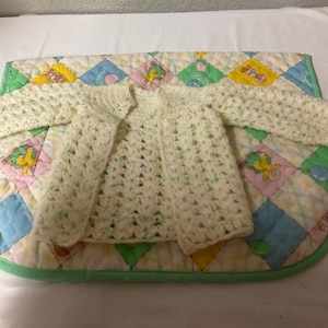 May include: A white crocheted baby cardigan with green flecks. The cardigan is laid out on a colorful quilted fabric.