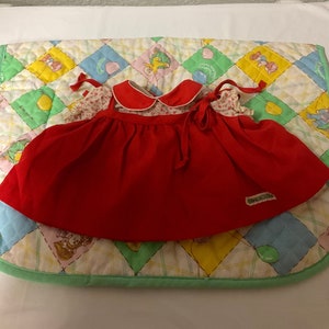 May include: Red and white doll's dress with a floral print and a red bow. The dress has a white collar with a red trim and the label "GUND" is visible on the bottom right corner.