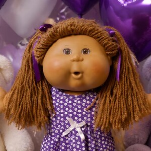 May include: A doll with brown yarn hair wearing a purple floral dress stands among several plush teddy bears in white and purple. The scene includes purple heart-shaped balloons, plates of chocolate chip cookies, brownies, and bowls of purple candies.