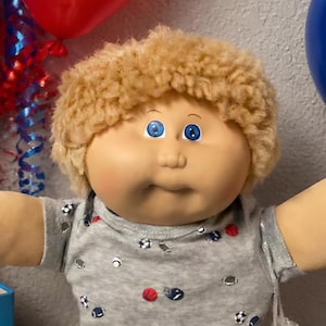 Op de afbeelding: Een Cabbage Patch Kids pop met krullend blond haar en blauwe ogen, gekleed in een grijs t-shirt met sportbalprint en een rode broek. De pop staat tussen speelgoed, waaronder twee kiepwagens, een rode auto en een bord met brownies.