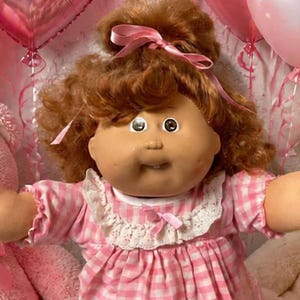 Peut inclure: Une poupée centrale aux cheveux bruns vêtue d'une robe à carreaux roses est entourée de deux poupées plus petites, de plusieurs ours en peluche roses et blancs, de ballons en forme de cœur, de cupcakes et de biscuits en forme de cœur.