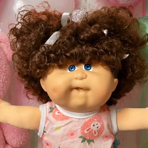 Peut inclure: Une poupée aux cheveux bruns bouclés et aux yeux bleus portant un haut sans manches à motif fraise, entourée d'ours en peluche, de deux petites poupées, d'une assiette de biscuits en forme de cœur, de brownies et de bonbons roses et verts.