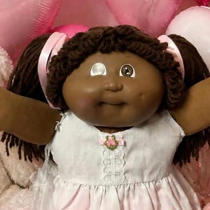 Könnte beinhalten: Eine dunkelhäutige Puppe mit braunen Wollhaaren in Zöpfen, bekleidet mit einem weiß-rosa Kleid mit Spitzenbesatz, umgeben von zwei kleineren Puppen, zwei Teddybären, rosa herzförmigen Luftballons, Cupcakes, herzförmigen Keksen und Süßigkeiten.