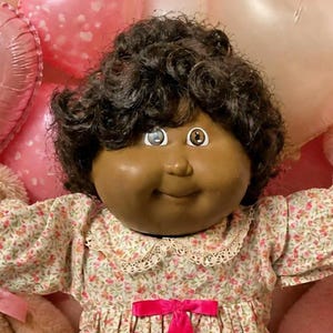 Peut inclure: Une poupée à la peau foncée aux cheveux noirs bouclés portant une robe à fleurs avec un ruban rose, des chaussettes blanches à volants et des chaussures blanches, entourée de deux petites poupées, deux ours en peluche et des brownies.