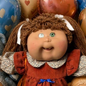 May include: A Cabbage Patch Kids doll with brown yarn hair, one white eye, and one green eye, wearing a rust-colored corduroy dress with a floral collar. Beside the doll are two smaller dolls in bonnets, a blue teddy bear, brownies, and cupcakes.