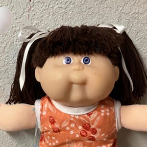 May include: A large doll with brown hair in pigtails, wearing an orange floral top and white lace pants, surrounded by four small baby dolls, a maroon teddy bear, a white teddy bear, scattered colorful candies, and maroon and white balloons.