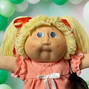 May include: A large blonde doll with blue eyes wearing a peach gingham dress with a white bow, surrounded by three smaller dolls, a green plush bear, colourful lollipops, assorted gummy sweets, and a balloon arch in shades of green and white.