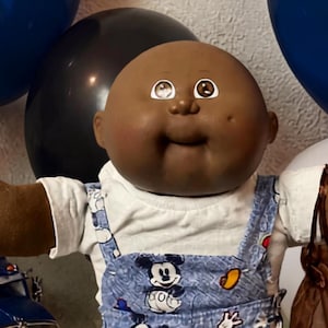 Op de afbeelding: Een donkerhuidige pop met een kaal hoofd draagt een wit shirt en een blauwe spijkerbroek met Mickey Mouse-print. De pop staat tussen verschillende speelgoedvoertuigen, waaronder een blauwe vrachtwagen, een zwarte trein en een rode raceauto.
