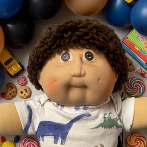 May include: A doll with brown yarn hair and dimpled cheeks wearing a white t-shirt with dinosaur prints and black pants, surrounded by blue and tan balloons, toy cars, stacks of books, chocolate chip cookies, lollipops, and a book titled The The Child Kid's Boook.