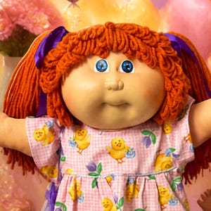 May include: A central doll with orange yarn hair in pigtails, wearing a pink gingham dress with yellow chick and purple flower prints, surrounded by four other dolls, heart-shaped balloons, cupcakes, and plates of colorful macarons.