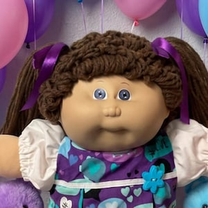 May include: A doll with brown yarn hair in pigtails, wearing a purple dress with heart and speech bubble patterns, white sleeves, and white shoes. The doll is flanked by a purple and a blue plush bear, surrounded by balloons and candy.