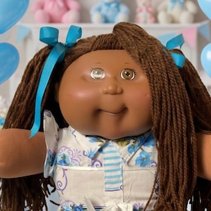 May include: A doll with dark brown yarn hair styled in pigtails with blue ribbons, wearing a white dress featuring a blue floral and striped pattern, white lace-trimmed socks, and white shoes, positioned among balloons and teddy bears.