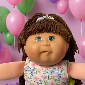 May include: A large doll with brown yarn hair and green eyes wearing a floral dress and light green pants, surrounded by smaller dolls, teddy bears, colorful balloons, cookies, and candies against a solid light purple background.