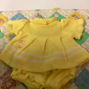 May include: A yellow knitted baby outfit, including a top and matching shorts. The top features a duck applique and white stripes. The shorts have a ruffled edge. The outfit is displayed on a colorful quilt.