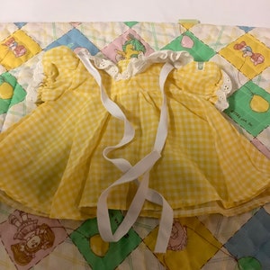 May include: A yellow and white gingham dress with white lace trim and white ribbon ties. The dress is on a quilted fabric with a yellow, green, and pink pattern.