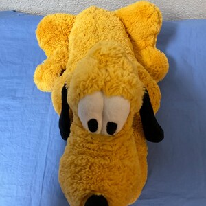 May include: A plush toy in the shape of a cartoon dog, primarily yellow with black ears and nose, and white eyes. The toy is lying down on a blue surface.