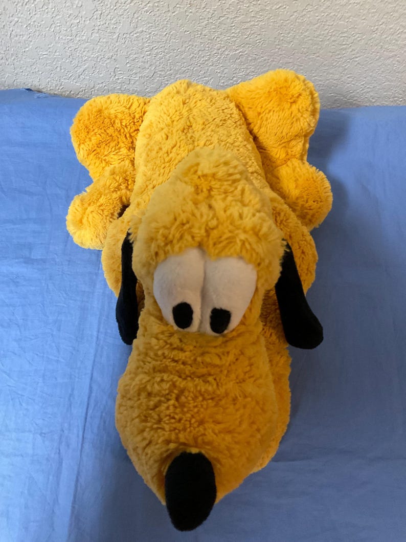 May include: A plush toy in the shape of a cartoon dog, primarily yellow with black ears and nose, and white eyes. The toy is lying down on a blue surface.