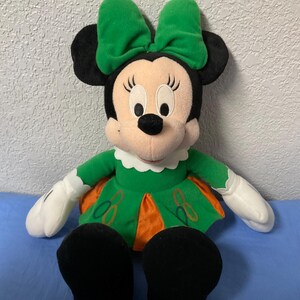 Puede incluir: Mu&ntilde;eca de peluche de Minnie Mouse con lazo verde, vestido y zapatos. La mu&ntilde;eca tiene orejas negras, cara rosa y guantes blancos. El vestido tiene detalles en naranja y bordados.