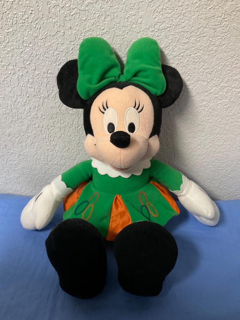 Puede incluir: Mu&ntilde;eca de peluche de Minnie Mouse con lazo verde, vestido y zapatos. La mu&ntilde;eca tiene orejas negras, cara rosa y guantes blancos. El vestido tiene detalles en naranja y bordados.