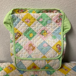 Puede incluir: Un portabebé acolchado verde con fondo blanco y un patrón de globos y muñecas coloridos. El portabebé tiene una solapa que se pliega y se asegura con un broche. El texto "Just grow in little things" está impreso en el portabebé.