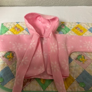 May include: A pink hooded fleece jacket with a white snowflake pattern. The jacket has a tie at the neck and long sleeves.