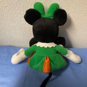 Puede incluir: Mu&ntilde;eca de peluche de Minnie Mouse, vista desde atr&aacute;s, con orejas y cabeza negras, un lazo verde y un vestido verde con detalles en naranja. La mu&ntilde;eca tiene guantes blancos y una cola negra.