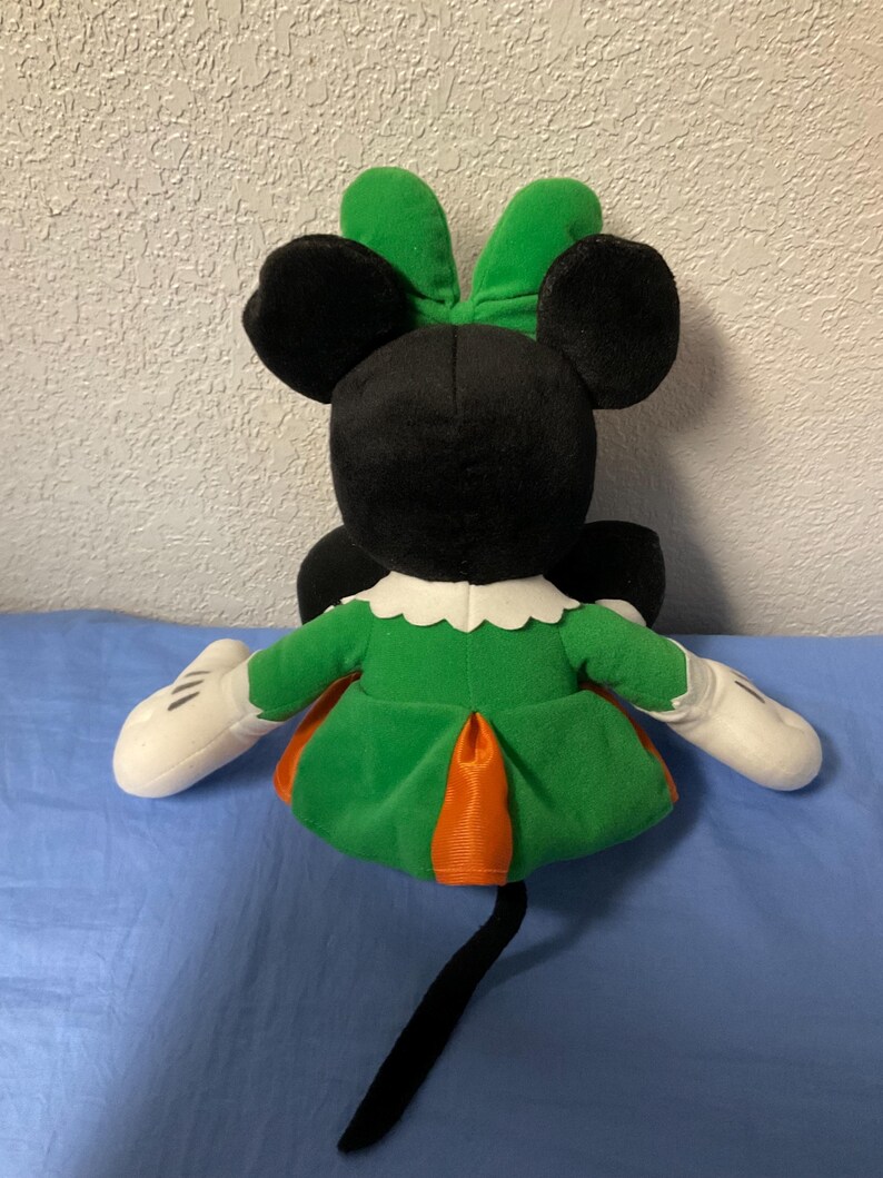 Puede incluir: Mu&ntilde;eca de peluche de Minnie Mouse, vista desde atr&aacute;s, con orejas y cabeza negras, un lazo verde y un vestido verde con detalles en naranja. La mu&ntilde;eca tiene guantes blancos y una cola negra.