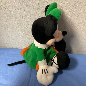 Puede incluir: Mu&ntilde;eca de peluche de Minnie Mouse con un vestido verde con ribete naranja, cuello blanco y lazo verde. La mu&ntilde;eca tiene orejas negras, cola negra y guantes blancos con detalles negros. La mu&ntilde;eca est&aacute; sentada sobre una superficie azul.