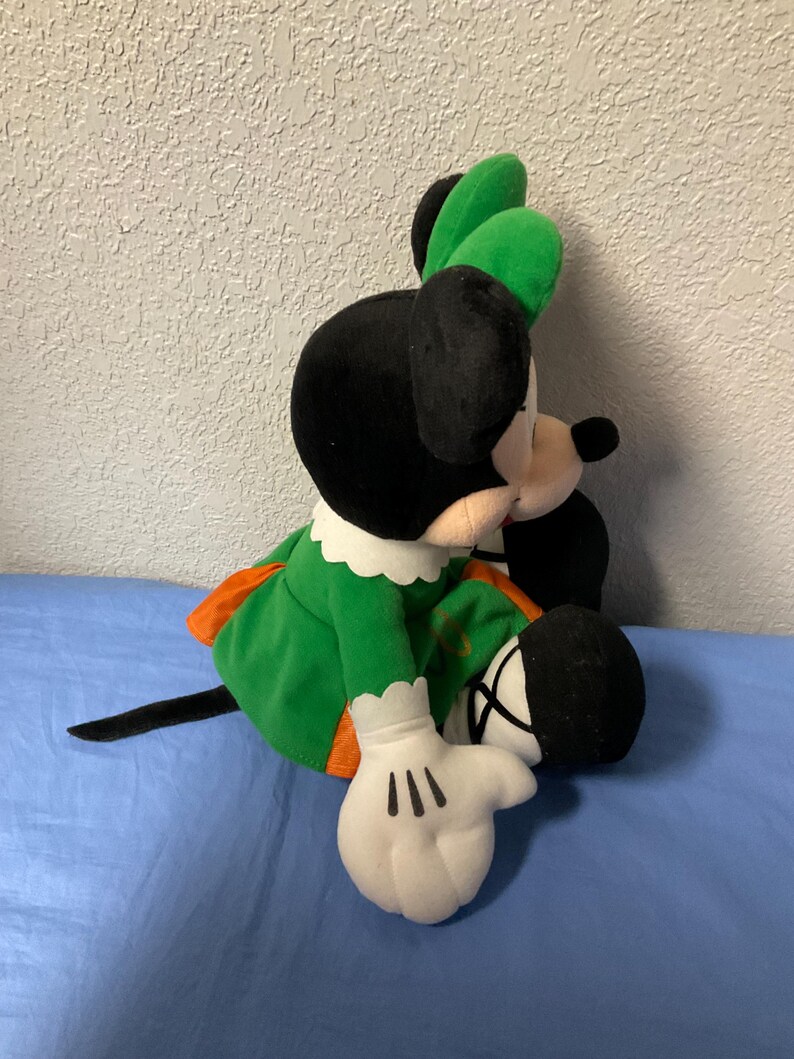 Puede incluir: Mu&ntilde;eca de peluche de Minnie Mouse con un vestido verde con ribete naranja, cuello blanco y lazo verde. La mu&ntilde;eca tiene orejas negras, cola negra y guantes blancos con detalles negros. La mu&ntilde;eca est&aacute; sentada sobre una superficie azul.