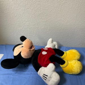 Pode incluir: Um grande brinquedo de pel&uacute;cia do Mickey Mouse est&aacute; deitado em uma superf&iacute;cie azul. O Mickey est&aacute; vestindo seus shorts vermelhos caracter&iacute;sticos com um &uacute;nico bot&atilde;o amarelo e luvas brancas. Ele tem orelhas pretas, um nariz preto e um grande sorriso.