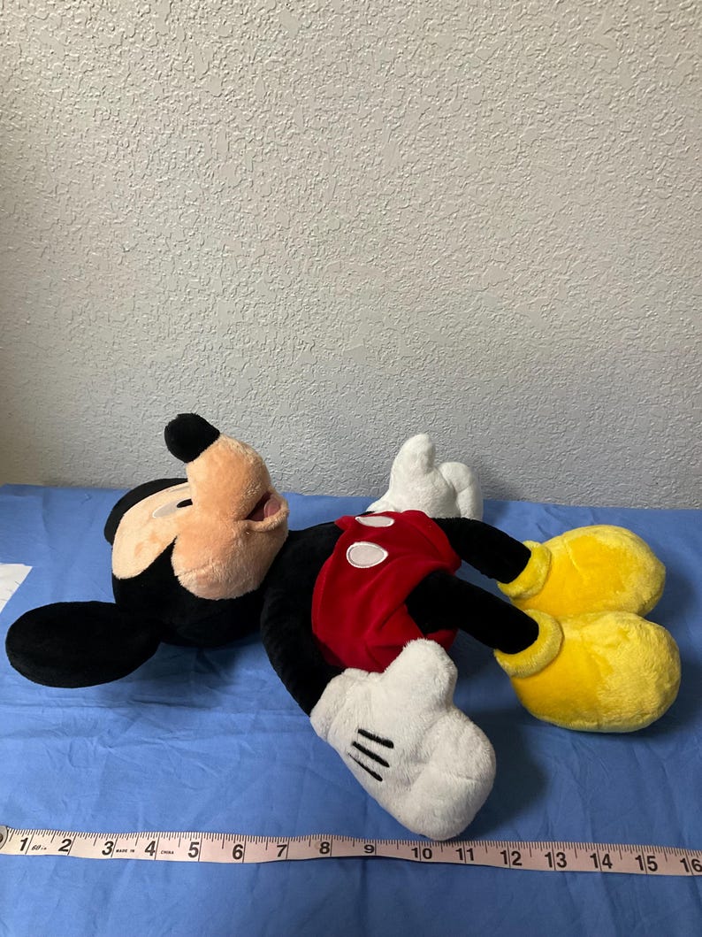 Pode incluir: Um grande brinquedo de pel&uacute;cia do Mickey Mouse est&aacute; deitado em uma superf&iacute;cie azul. O Mickey est&aacute; vestindo seus shorts vermelhos caracter&iacute;sticos com um &uacute;nico bot&atilde;o amarelo e luvas brancas. Ele tem orelhas pretas, um nariz preto e um grande sorriso.