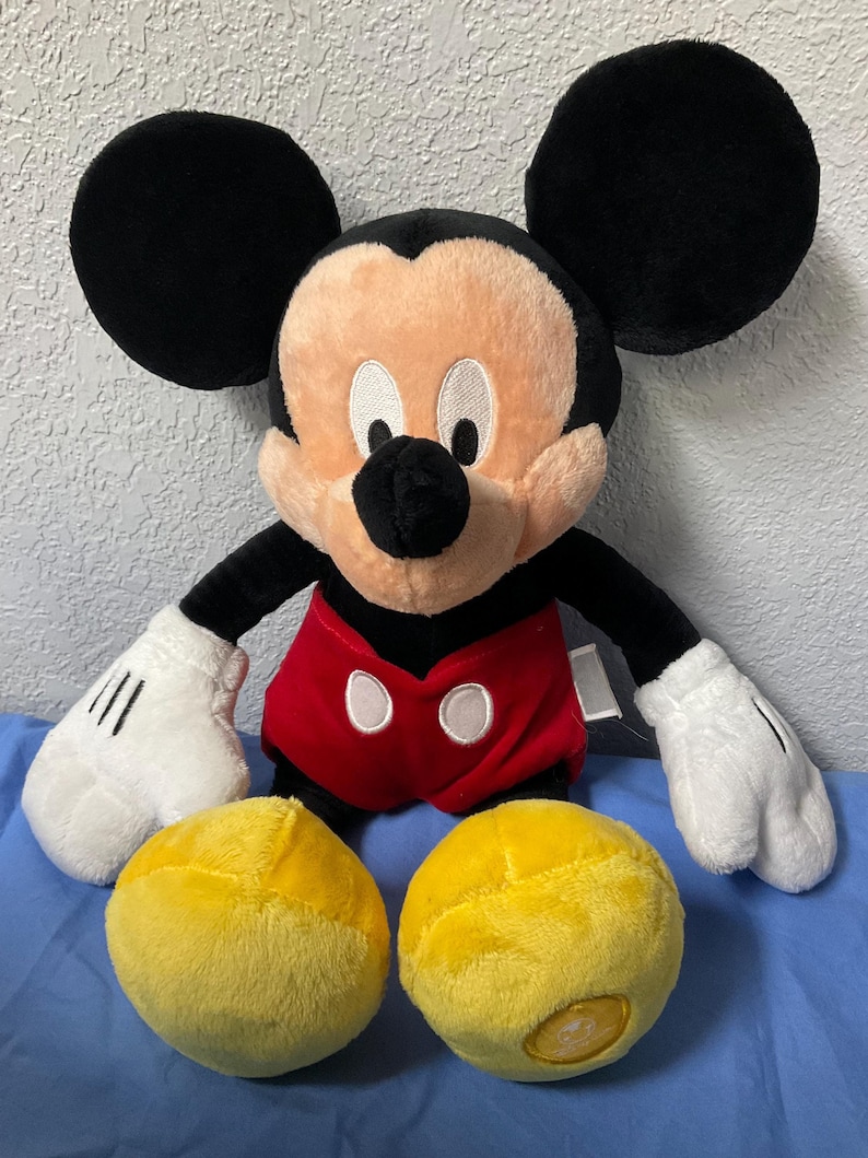 Pode incluir: Um grande brinquedo de pel&uacute;cia do Mickey Mouse com orelhas pretas, uma camisa vermelha e branca e sapatos amarelos. O brinquedo est&aacute; sentado em uma superf&iacute;cie azul.