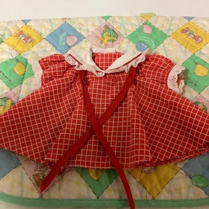 Puede incluir: Vestido de cuadros rojos y blancos con ribete de encaje blanco y un lazo rojo. El vestido está sobre un fondo de tela acolchada de colores.