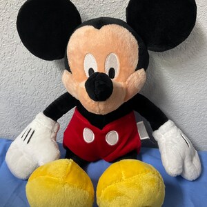 Pode incluir: Um brinquedo de pel&uacute;cia do Mickey Mouse com orelhas pretas, uma camisa vermelha com bot&otilde;es brancos e sapatos amarelos. O brinquedo est&aacute; sentado em uma superf&iacute;cie azul.