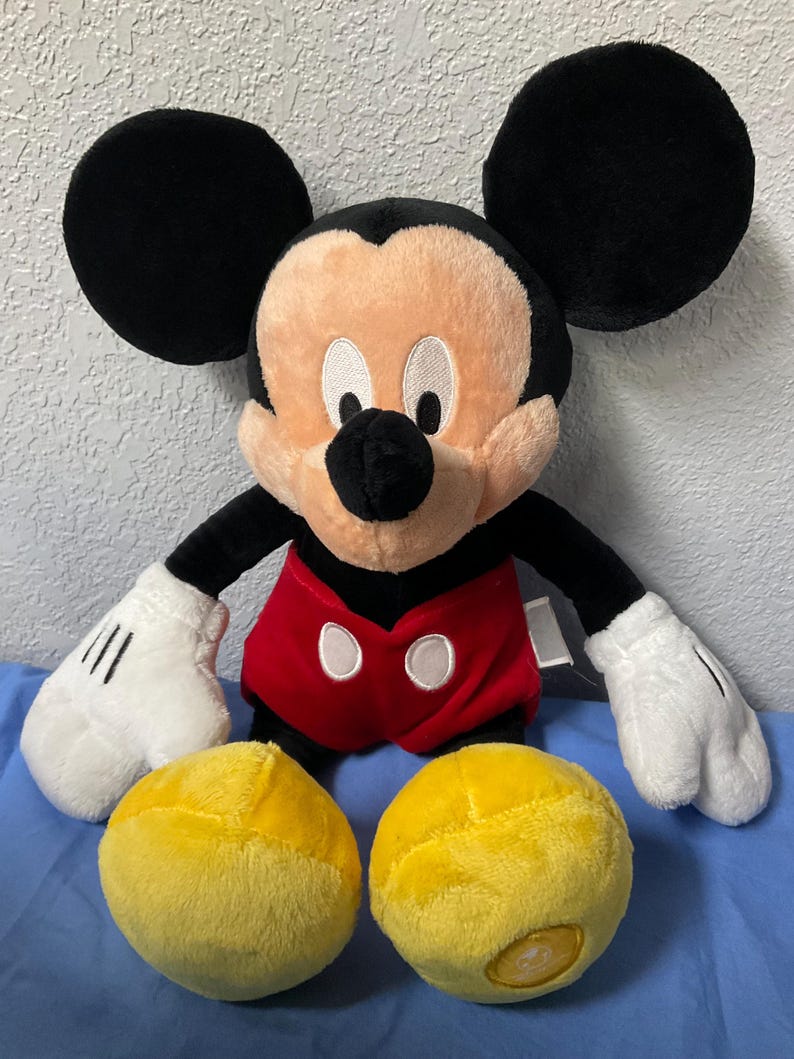 Pode incluir: Um brinquedo de pel&uacute;cia do Mickey Mouse com orelhas pretas, uma camisa vermelha com bot&otilde;es brancos e sapatos amarelos. O brinquedo est&aacute; sentado em uma superf&iacute;cie azul.