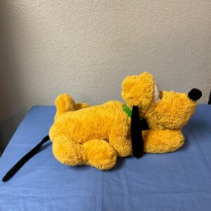 May include: A plush, yellow dog toy with a black tail and ears, and a green collar. The dog has a black nose and white eyes. The toy is lying on a blue surface.