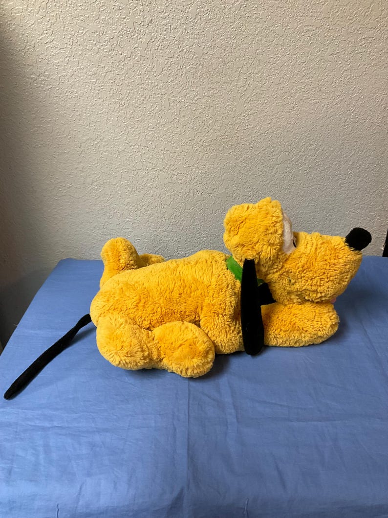 May include: A plush, yellow dog toy with a black tail and ears, and a green collar. The dog has a black nose and white eyes. The toy is lying on a blue surface.