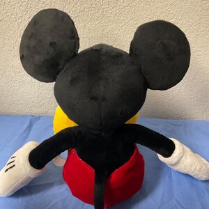 Pode incluir: Um brinquedo de pel&uacute;cia Mickey Mouse preto e vermelho. O brinquedo est&aacute; sentado em uma superf&iacute;cie amarela. O brinquedo tem grandes orelhas pretas, luvas brancas e uma cauda vermelha.
