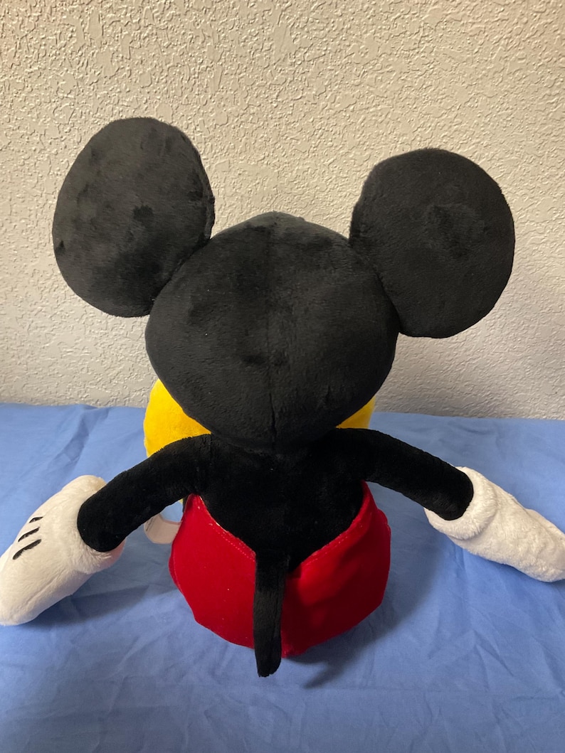 Pode incluir: Um brinquedo de pel&uacute;cia Mickey Mouse preto e vermelho. O brinquedo est&aacute; sentado em uma superf&iacute;cie amarela. O brinquedo tem grandes orelhas pretas, luvas brancas e uma cauda vermelha.
