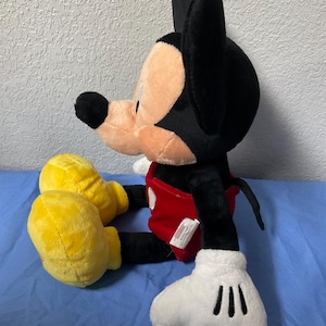 Pode incluir: Um grande brinquedo de pel&uacute;cia do Mickey Mouse sentado com as pernas cruzadas. Ele est&aacute; vestindo shorts vermelhos com um bot&atilde;o branco e sapatos amarelos. Suas orelhas s&atilde;o pretas e suas luvas s&atilde;o brancas.