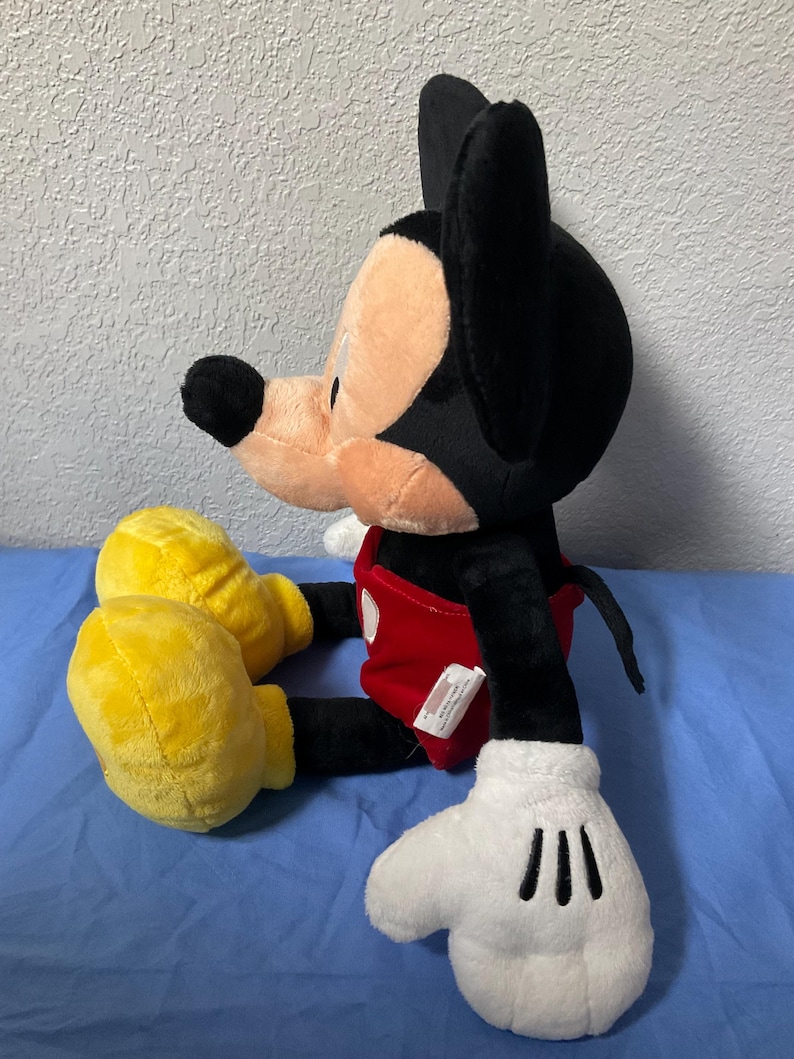 Pode incluir: Um grande brinquedo de pel&uacute;cia do Mickey Mouse sentado com as pernas cruzadas. Ele est&aacute; vestindo shorts vermelhos com um bot&atilde;o branco e sapatos amarelos. Suas orelhas s&atilde;o pretas e suas luvas s&atilde;o brancas.