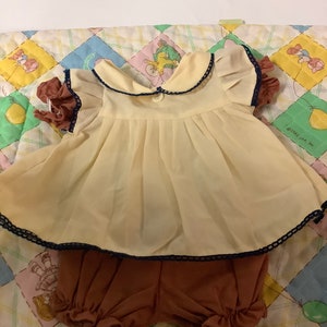 Puede incluir: Un vestido amarillo con ribete azul y pantalones cortos marrones con volantes. El vestido tiene un botón blanco y un ribete de encaje blanco. El conjunto está sobre una manta acolchada con un patrón colorido.