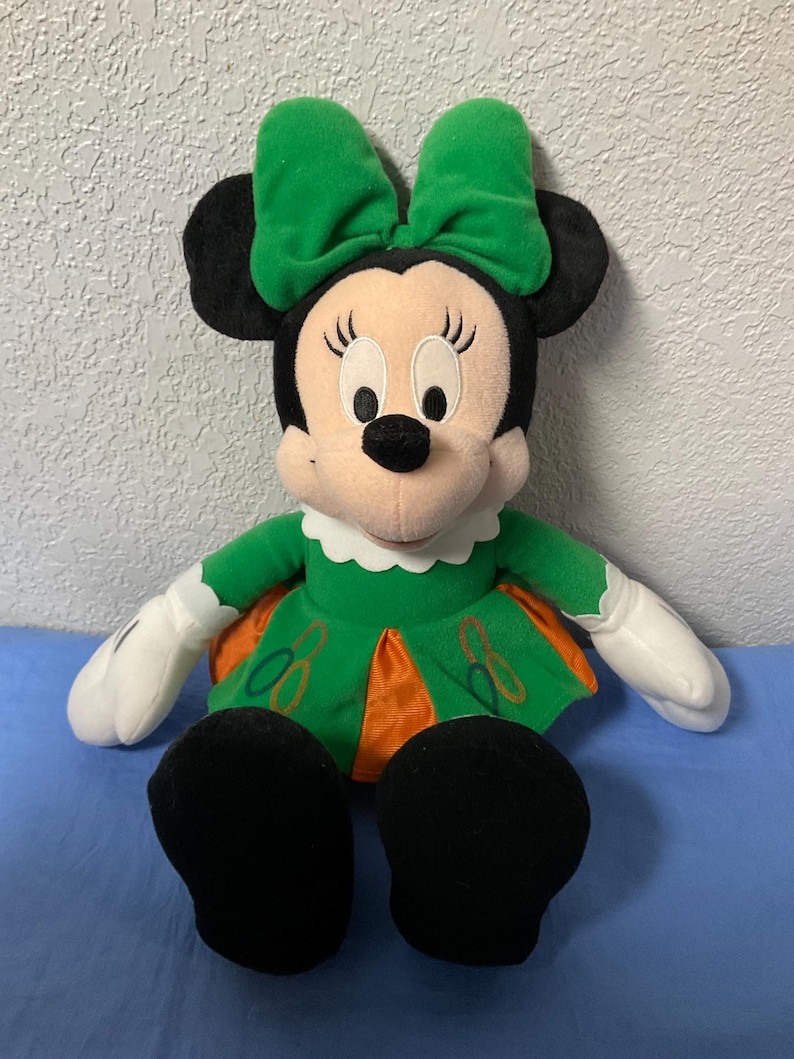 Puede incluir: Mu&ntilde;eca de peluche de Minnie Mouse con un vestido verde con detalles en naranja y un lazo verde a juego. La mu&ntilde;eca tiene orejas negras, nariz negra y guantes blancos. La mu&ntilde;eca est&aacute; sentada sobre una superficie azul.
