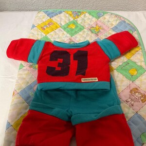 May include: A red and teal doll outfit with the number 31 on the front. The outfit includes a long-sleeved shirt and shorts. The outfit is displayed on a colourful patchwork quilt.