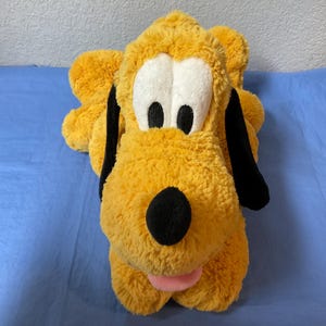 May include: A plush toy of Pluto, a yellow dog with black ears and a black nose. The dog has large white eyes with black pupils and a pink tongue. The toy is lying on a blue surface.