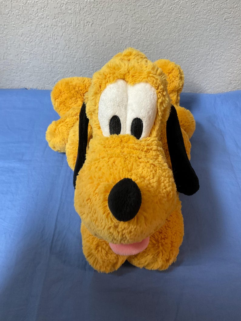 May include: A plush toy of Pluto, a yellow dog with black ears and a black nose. The dog has large white eyes with black pupils and a pink tongue. The toy is lying on a blue surface.