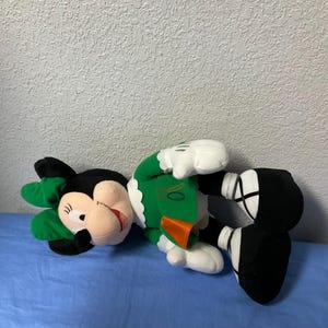 Puede incluir: Mu&ntilde;eca de peluche de Minnie Mouse con un atuendo verde con cuello blanco y lazo verde a juego. La mu&ntilde;eca tiene orejas negras, boca roja y guantes y zapatos blancos. La mu&ntilde;eca est&aacute; tumbada sobre una superficie azul.